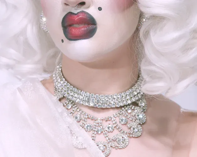 Close crop colour photo of a glamorously made-up person. White face-powder, full-red and black lipstick, beauty spots, sliver necklace and white blond hair