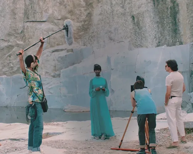 Film still. A film crew circle a Black woman in a long blue dress. They are shooting outdoors in a quarry