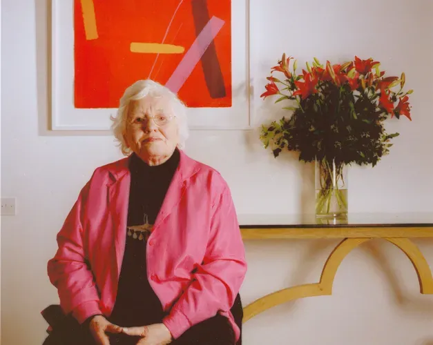 An older woman with white hair sits indoors wearing a bright pink jacket, with an abstract red and orange artwork framed on the wall behind her and a vase of red lilies on a table to her side.