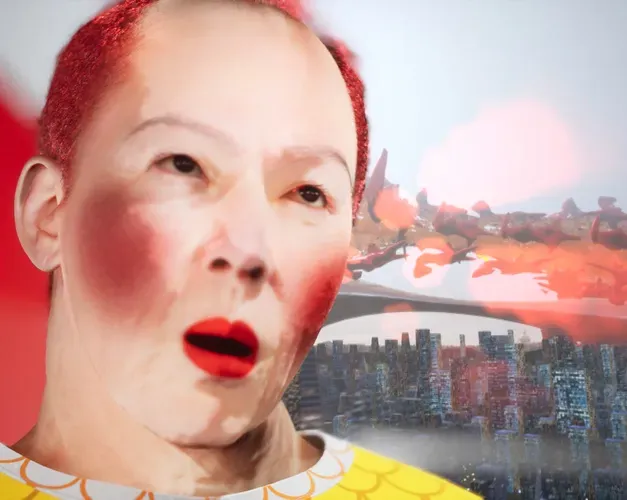 Digital video still. A close-up CGI rendering of an asian man with white make-up on his face, rouge on his cheeks and dyed short red hair. Over his should we can see two types of landscape. Rows of skyscrappers and above this red amorphous shapes floating in a digital pink hued sky.