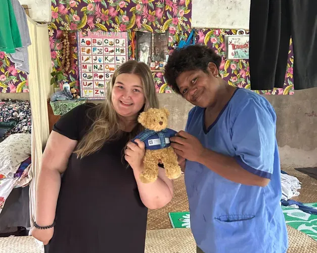 Two people standing indoors on a woven mat, smiling and holding a University of Dundee teddy bear together. Colourful fabric and hanging clothes are visible in the background.