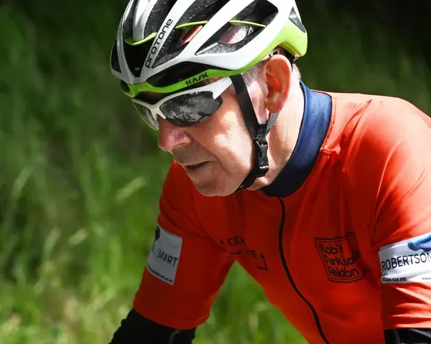 A close-up of a cyclist wearing a white and green helmet, sunglasses, and an orange jersey.
