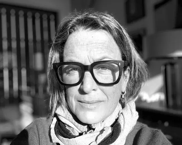 A black-and-white close-up portrait of a person wearing large square glasses, looking toward the camera with bright sunlight casting strong shadows across their face.