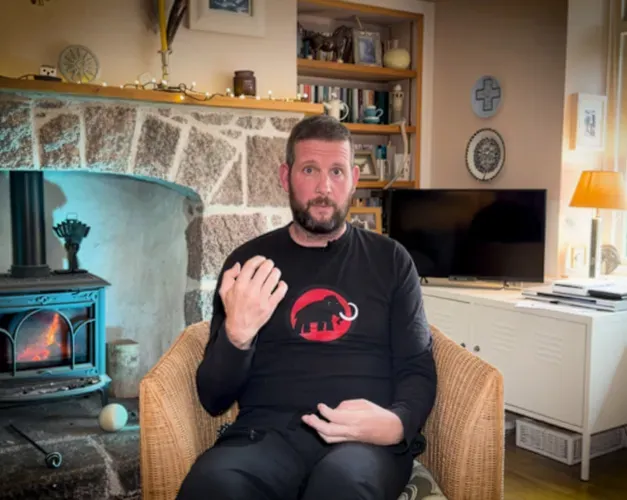 man sitting on sofa, holding hands up, in living room with wood burning fire behind