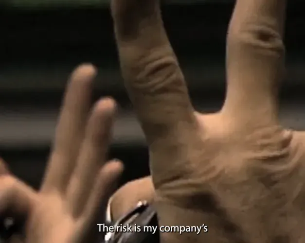 film still. Close up of fingers and hangs held high in the air with caption 'the risk is my company's'