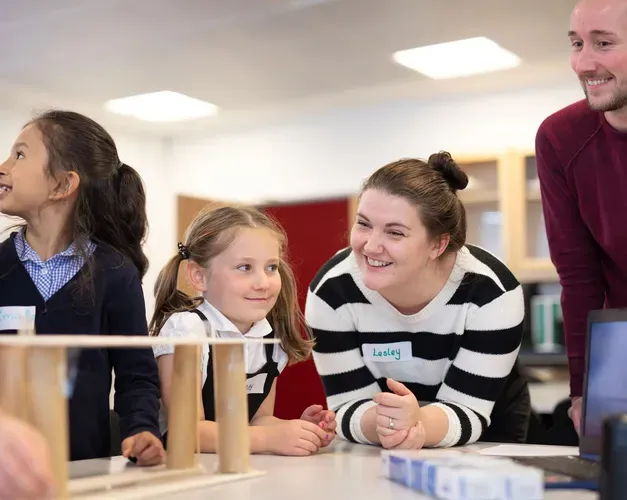 Two adults lean over a table smiling as young children in school uniforms observe and interact with a simple wooden construction during a classroom activity