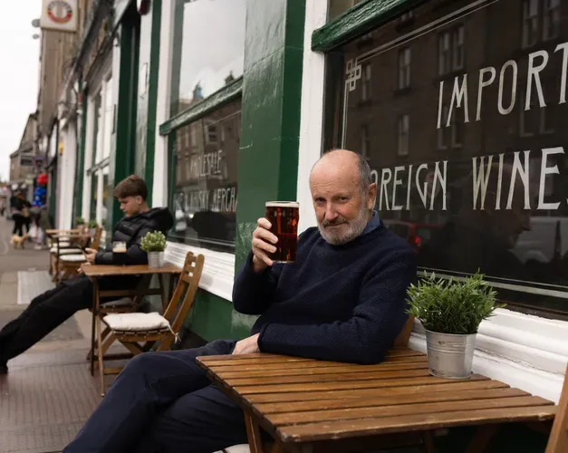 A smiling Fred Macaulay sits at a bistro-style table outside the Speedwell Bar - better-known as Mennies - on Perth Road in Dundee, Scotland. He holds a pint of beer up in a "cheers" gesture