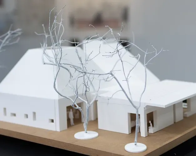 miniature model of a large house made from white material. Tiny little figures stand nearby and there are small branches outside to resemble trees in the winter without their leaves