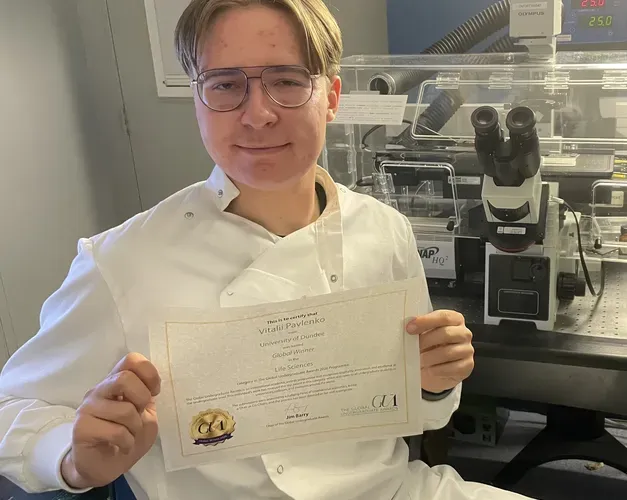 A man wearing a lab coat sat in front of a confocal microscope holding a certificate