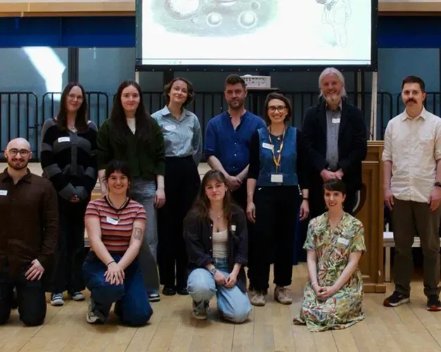 A group of 12 adults pose for a photograph in two rows in a university lecture hall, with several people kneeling in front and others standing behind. A projection screen hangs above them
