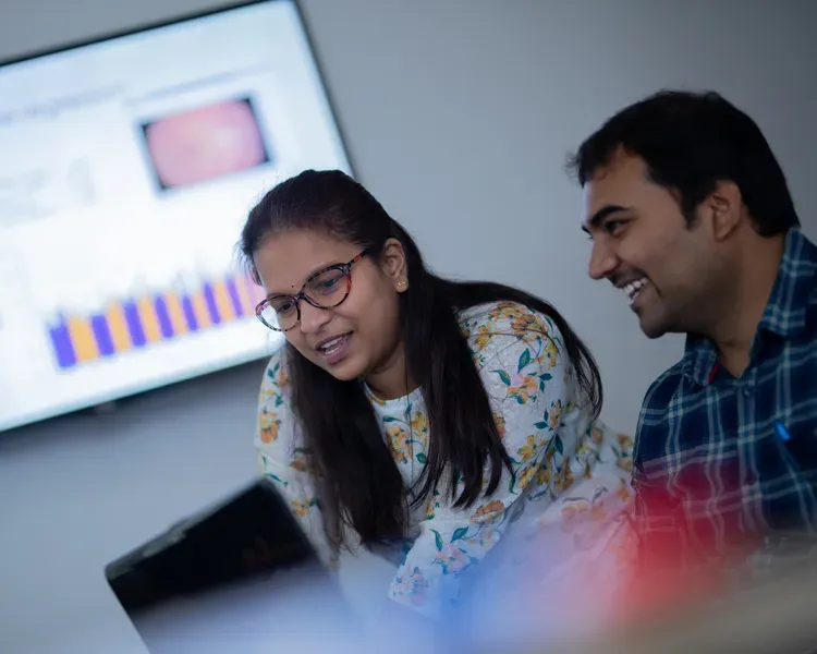 Students compare data on a laptop with a large screen behind them showing charts