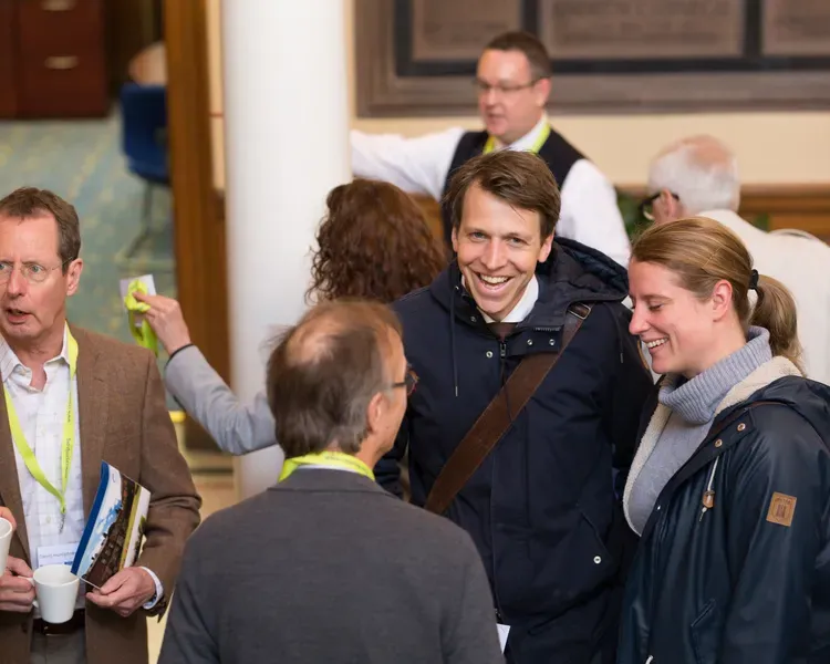 A group of people standing at a conferences smiling and laughing. Some are holding cups and brochures