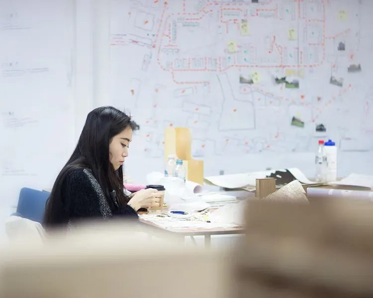 student working at a table in the architecture studio with plans pinned up on the walls