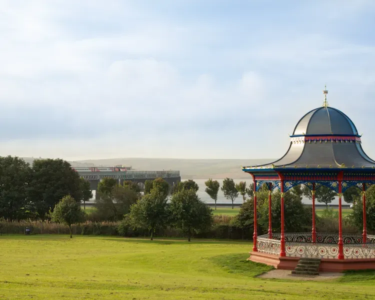 An LNER branded HST with 9 coaches crosses the Tay Bridge with a park and bandstand in the foreground