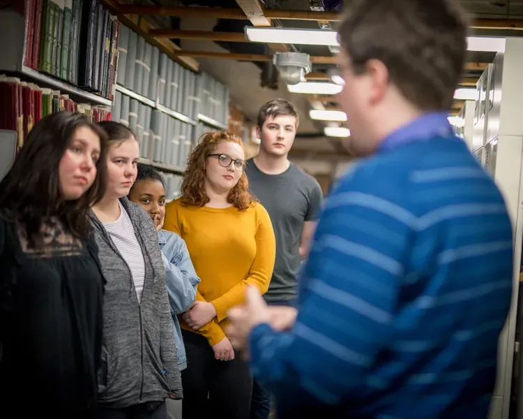 A group of students listen to a man talking while standing between archive shelving