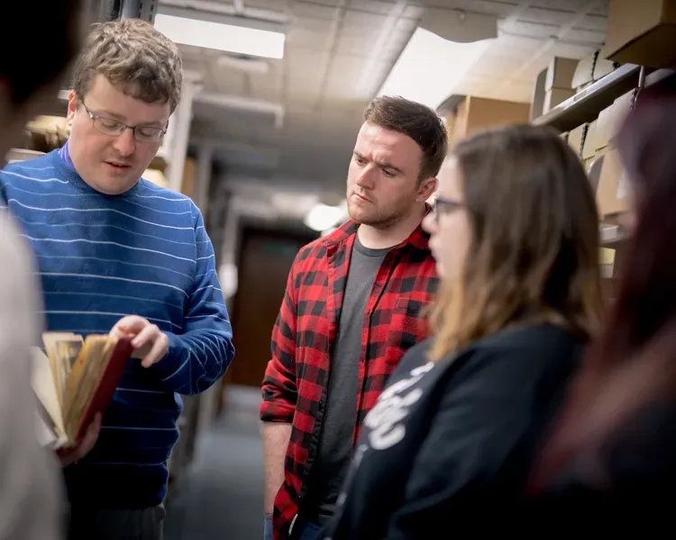 A group of students standing in an archive listening to a man holding an old book