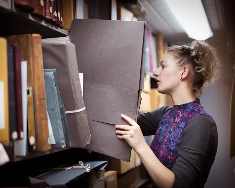 A student places a large folder back on archive shelving
