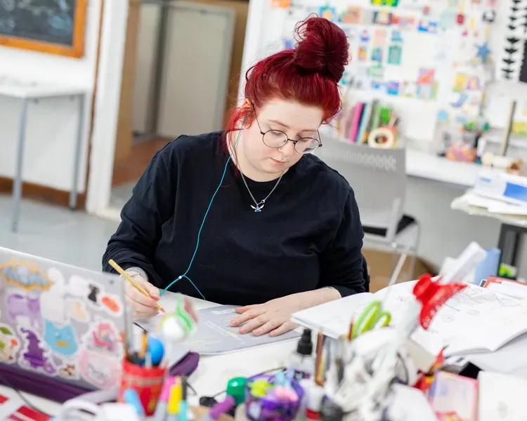 female student working in illustration studio with laptop and colourful materials