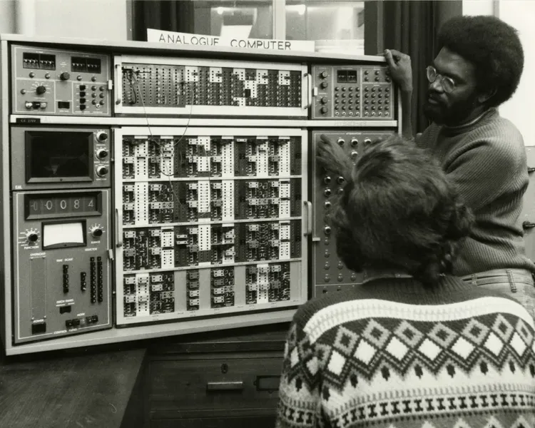 Two men work on an analogue computer, with dials, and wire terminals
