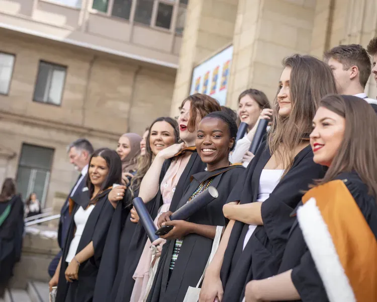 Graduates with certificats and wearing full roves standing on steps and smiling to have their photo taken