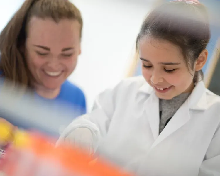 A child wearing a white labcoat with a woman wearing a blue teeshirt, both smiling