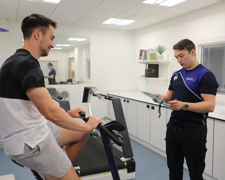 Man sits on exercise bike while another looks on and takes notes.