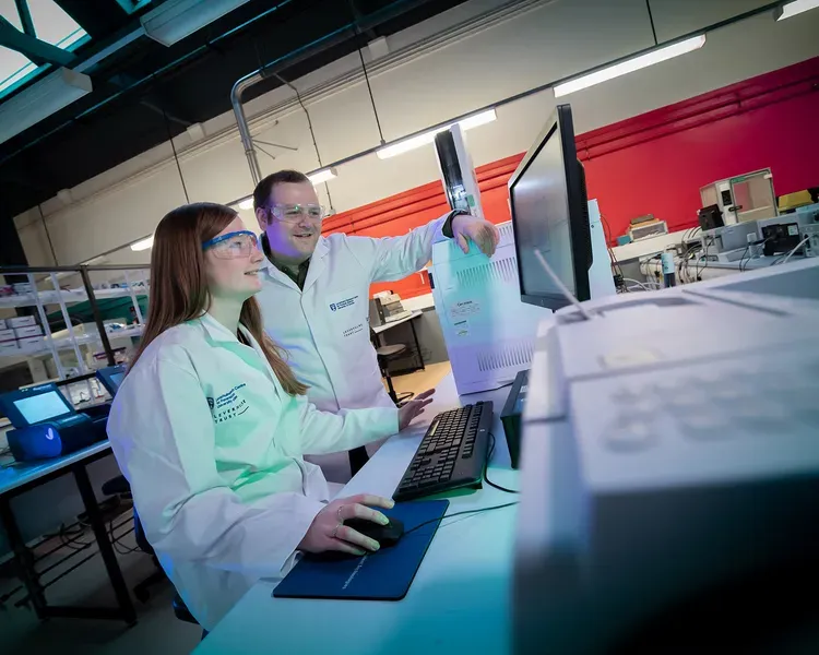 Two forensic scientists in lab equipment smiling as they view a computer screen.