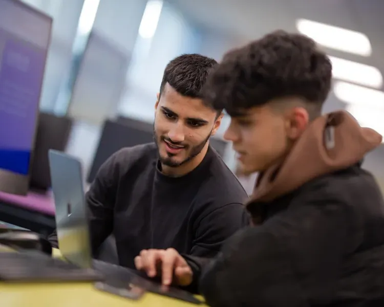 two students work on a laptop together, other computers are in the background along with large monitors