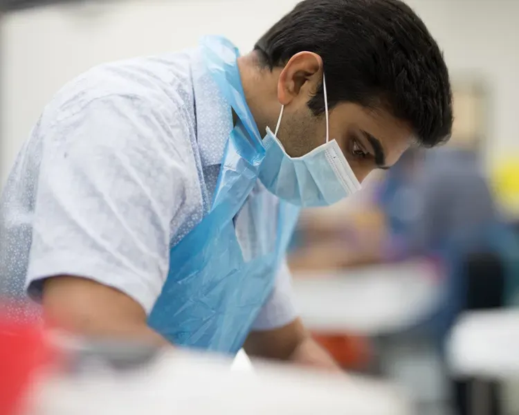 Orthopaedic student wearing a medical mask and gown. He is looking down at something he is operating on outside of the photo.  