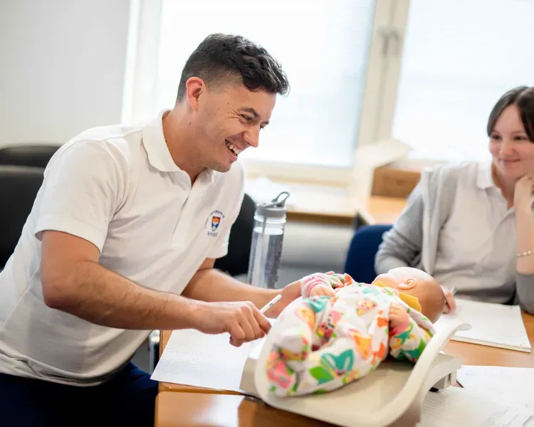 A male and female, both in Dundee University branded shirts are sitting at a table with a baby doll between them.