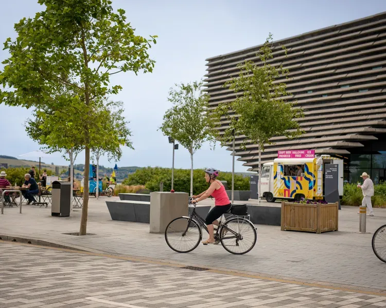 People sat a picnic tables outside V&A Dundee as cyclists go past