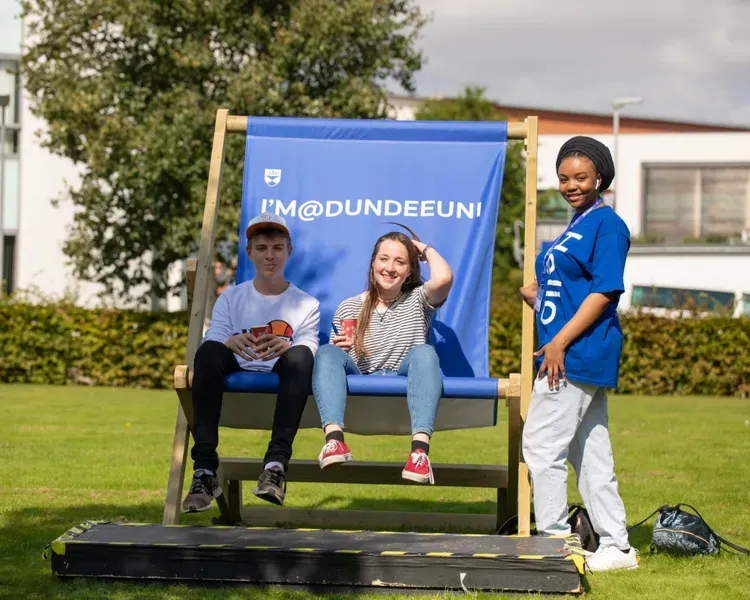 Three young adults sitting together on a University of Dundee branded lawn chair, enjoying leisure time in a grassy area under clear skies.