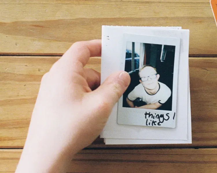 A hand holds a stack of cards with a polaroid photo on top against a wooden background. In the upper right hand corner is an orange card with a number of short lists typed on it. There are tickboxes against each item on the lists
