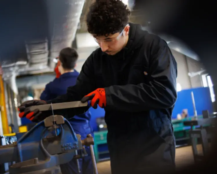 A student in a boiler suit and gloves works a piece of metal being held in a vice