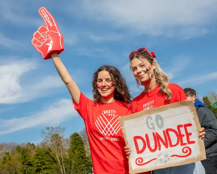 Two students in red t-shirts hold up a red foam finger and a cardboard sign reading Go Dundee as they support varsity sports teams at the University's Riverside playing fields