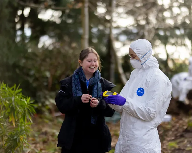 One person in white ppe and a face mask holds a yellow device showing it to someone, there are trees around them.