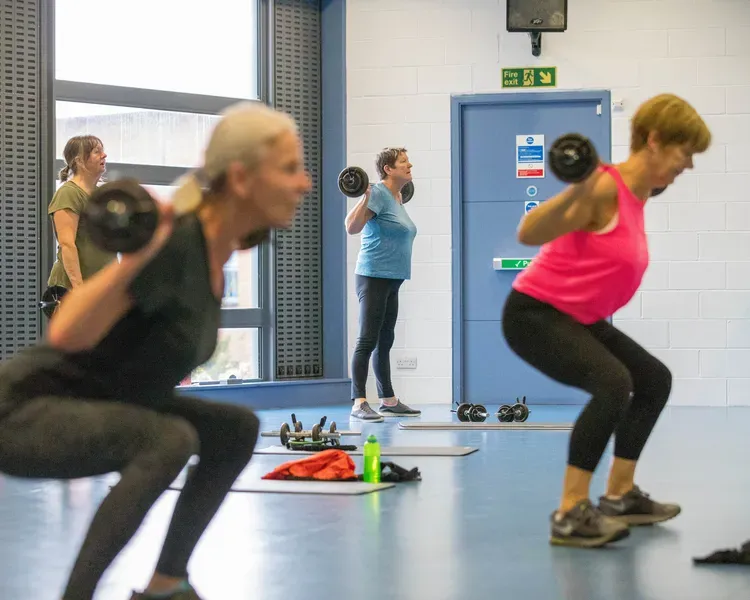 A group of women are in a room squatting while lifting weights on bars across their shoulders.