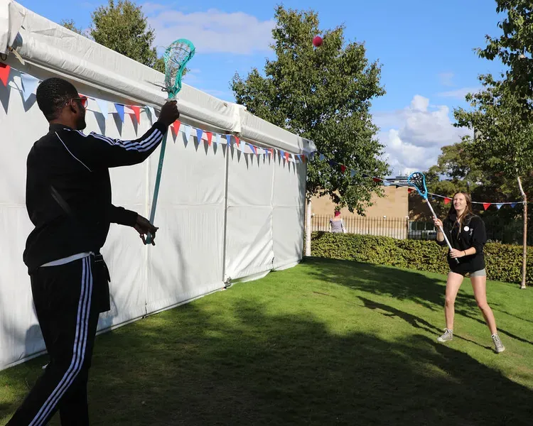 Two people stand outside of a marquee playing with lacrosse sticks.