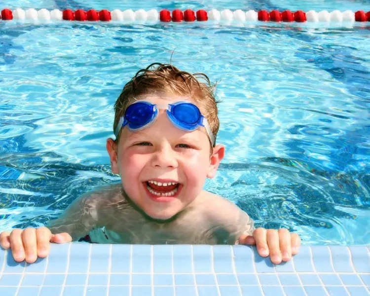 A child in a swimming pool with swimming goggles on his head holding onto the edge of the pool