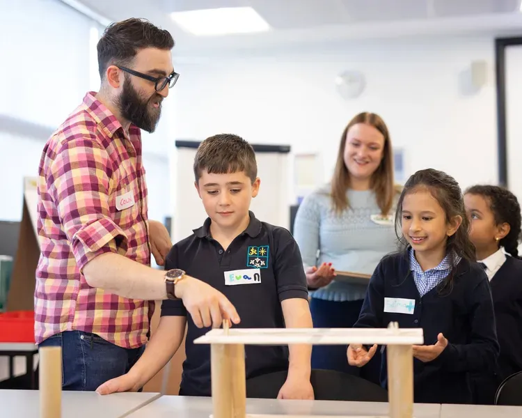 A teacher wearing glasses and a plaid shirt guides a group of schoolchildren as they build a small wooden structure on a table in a bright classroom