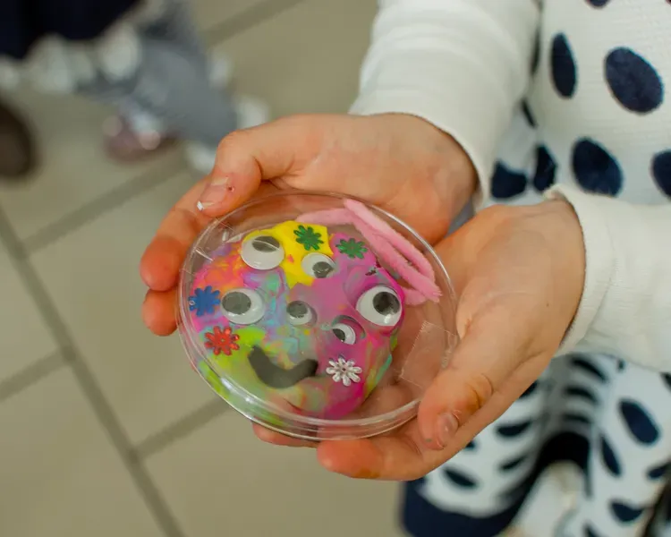 Child holding a petri dish with a colorful handmade microbe craft decorated with googly eyes, pipe cleaners, and foam flowers.