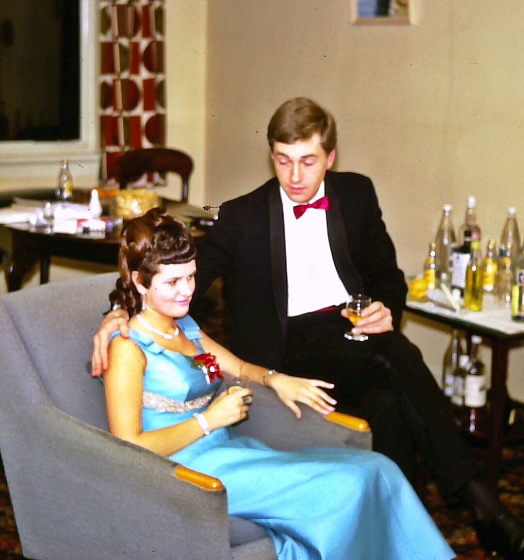 a man sitting on the arm of a chair with his arm around his wife who is sitting in the chair