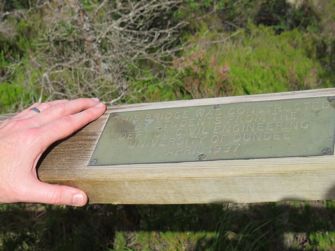 A man's hand, a silver wedding band on the ring finger, rests on the bridge handrail. Next to it, a faintly weathered metal plaque is screwed down. It reads "This bridge was constructed by students from the Dept of Civil Engineering University of Dundee April 1997" in block capitals.