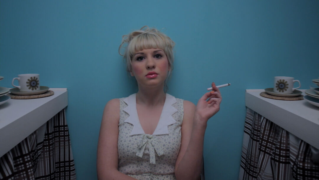 Film still from ‘Egg and Fag’. Actress sits in a kitchen facing camera holding a cigarette