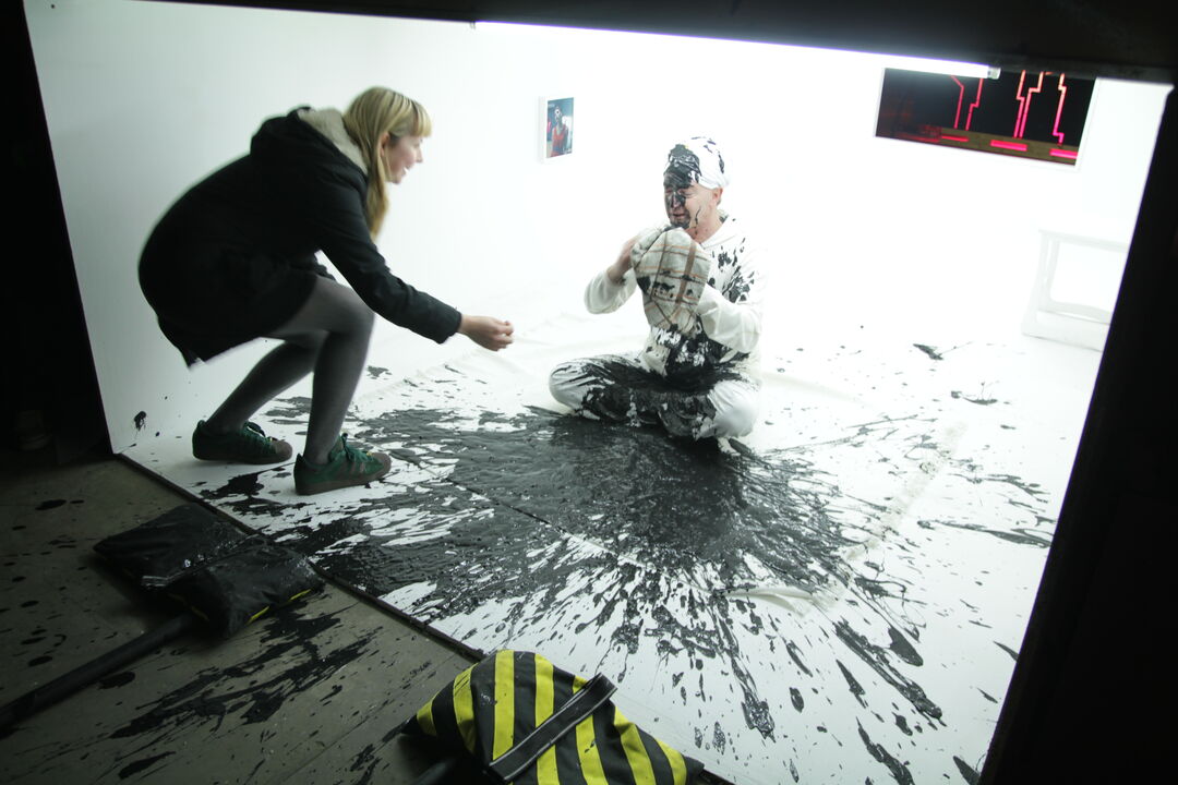Behind the scenes photo from a film set at Tin Roof. Actor sits on floor with paint splashed on and around him while Rose Hendry directs.