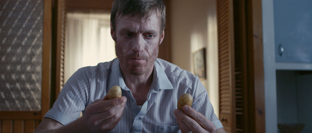Film still from ‘Stovies’. Actor Doug Russell sits in kitchen holding two small potatoes