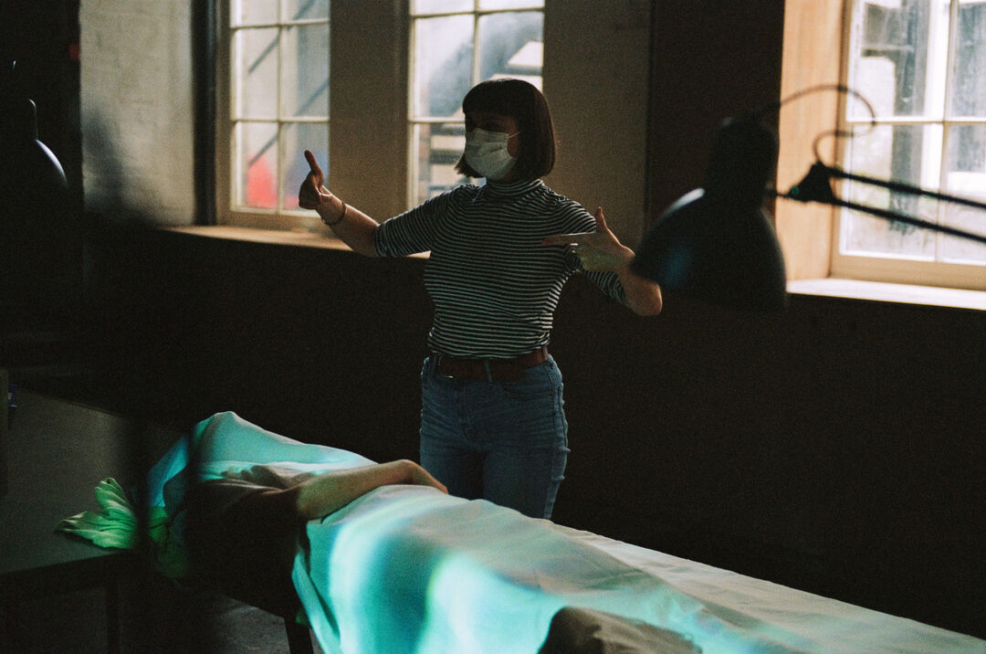 Rose Hendry behind the scenes for The Dead Are Jewels To Me, wearing a face mask, directing a scene with an actor playing a deceased individual