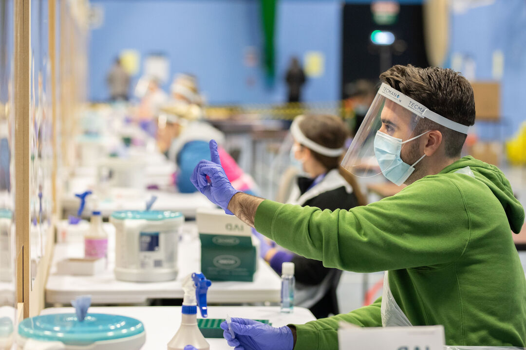 A person sitting in a Covid testing centre, wearing a mask and gloves, holding a syringe