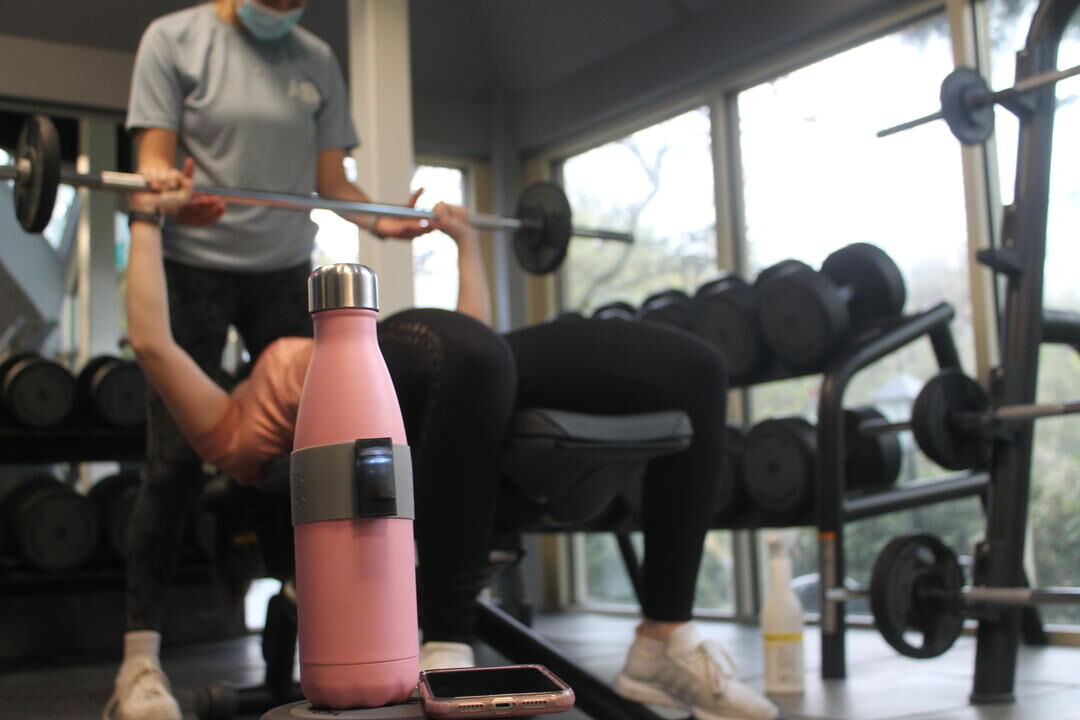 A student helps another student to use a gym's bench press. In the foreground, there is a sports bottle with a special band created as part of a student project called Mygym.