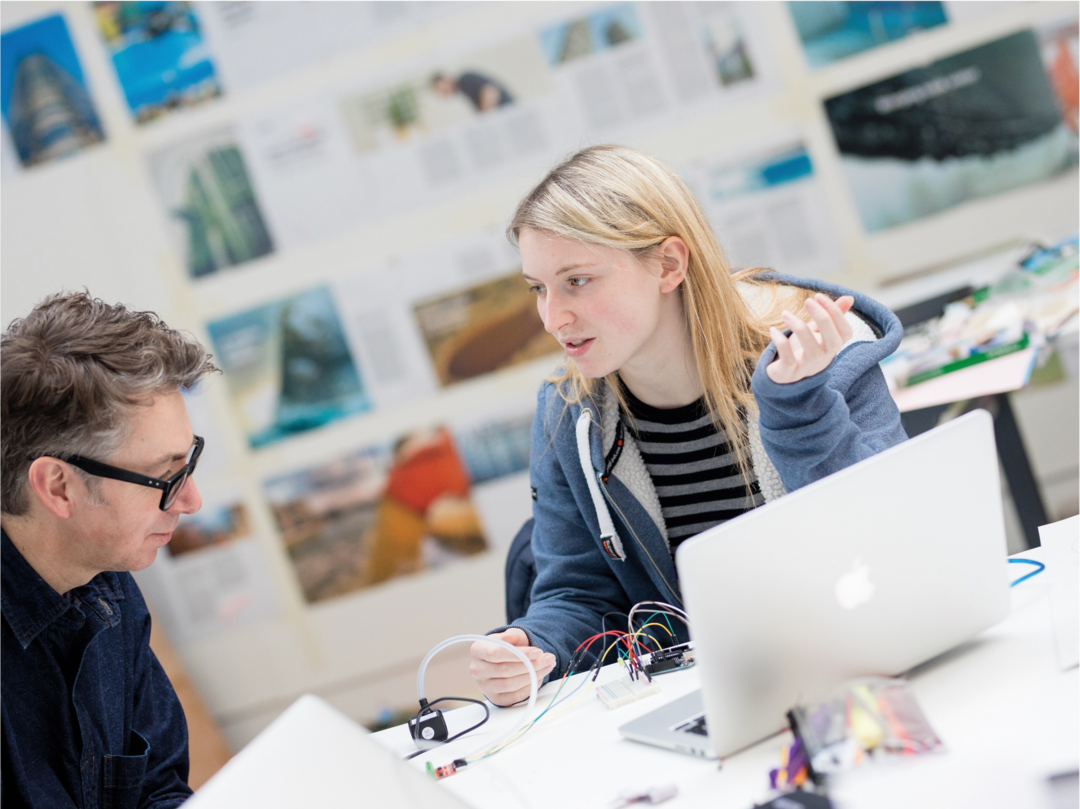 A female student speaking to a male lecturer in a studio environment with small electronic components and a laptop on the table in front of them.